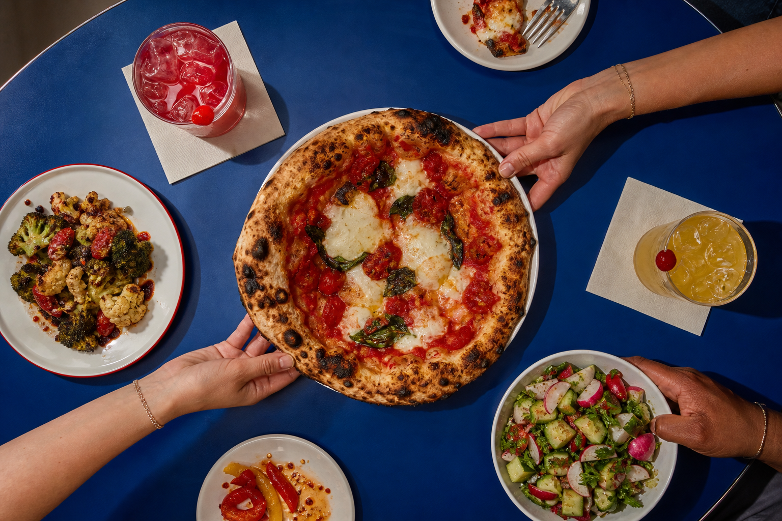 Overhead view of blistered flatbread, chopped salad, roasted vegetables, and two colorful zero-proof drinks on a cobalt table, with hands reaching in from the edges.
